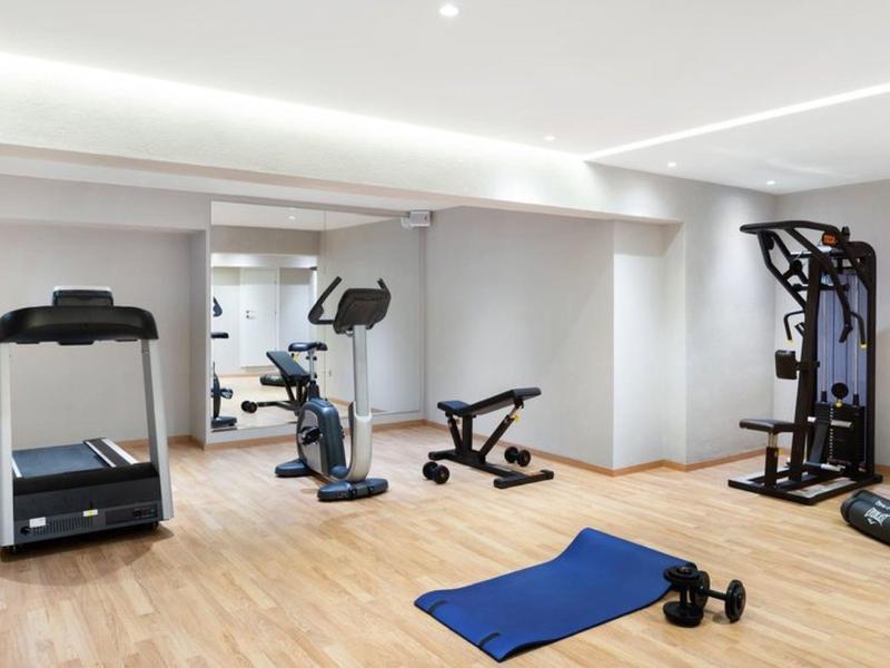Salle de sport d'hôtel moderne avec tapis roulant, vélo d'appartement, poids et tapis d'exercice bleu sur sol en bois.