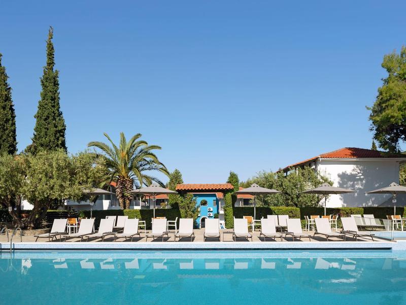 Piscine extérieure avec chaises longues et arbres sous un ciel bleu clair dans un complexe.