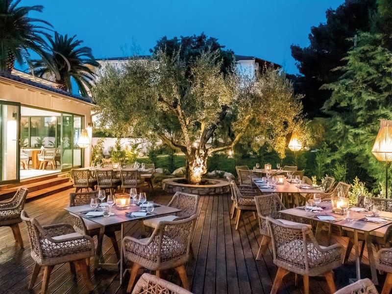 Restaurant extérieur d'hôtel avec chaises en osier, terrasse en bois, éclairage décoratif et verdure luxuriante au crépuscule.