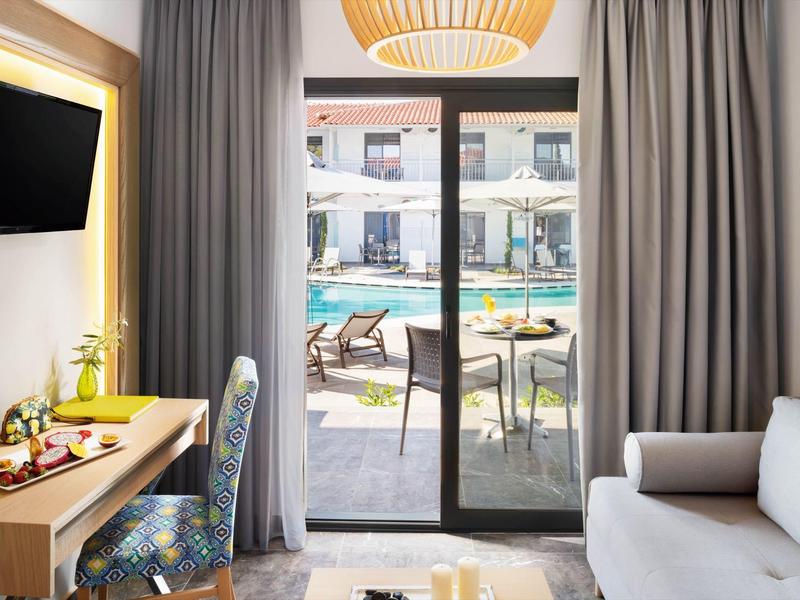 Chambre d'hôtel avec balcon donnant sur la piscine, mobilier moderne et rideaux gris.