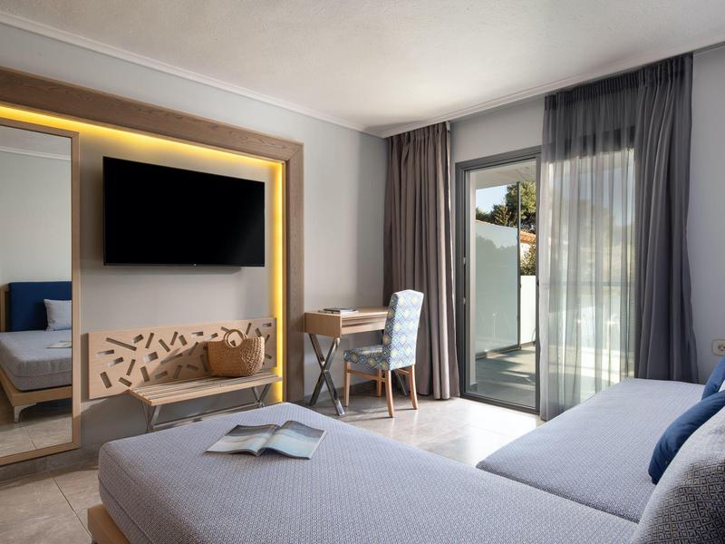 Chambre d'hôtel moderne avec lit, TV, bureau, chaise et porte-fenêtre vers balcon.