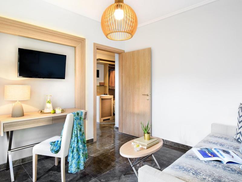 Chambre d'hôtel lumineuse avec bureau, chaise, TV murale, canapé, table ronde et porte ouverte.