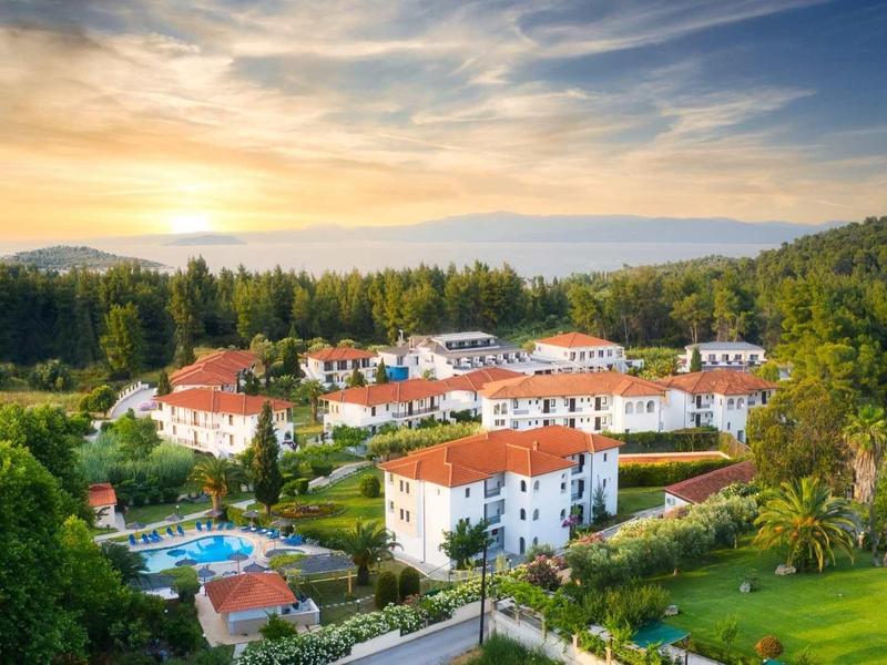 Resort con edificios blancos y techos rojos rodeado de árboles y una piscina bajo un cielo al atardecer