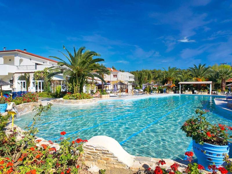 Piscina grande rodeada de flores y palmeras en un hotel bajo cielo azul despejado.