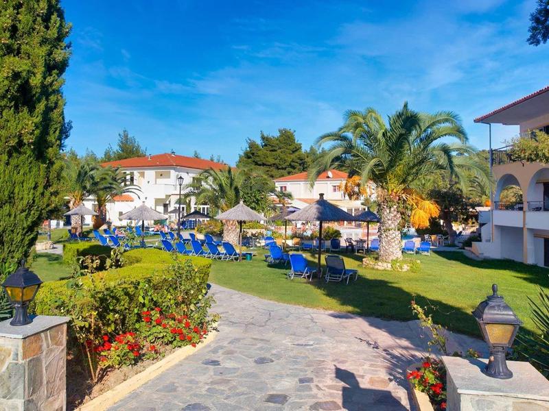 Amplio complejo hotelero con camino, jardín, palmeras y tumbonas azules bajo cielo azul.