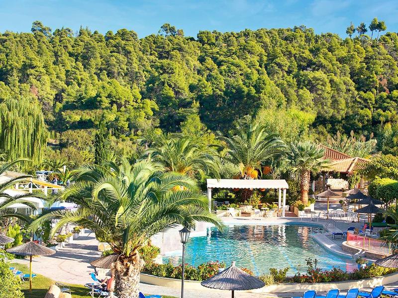 Piscina de hotel con tumbonas y palmeras frente a colina boscosa bajo clima soleado.