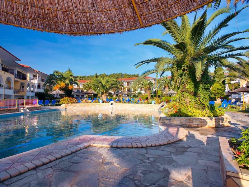 Área de piscina del hotel con palmeras, tumbonas y apartamentos alrededor bajo cielo despejado.