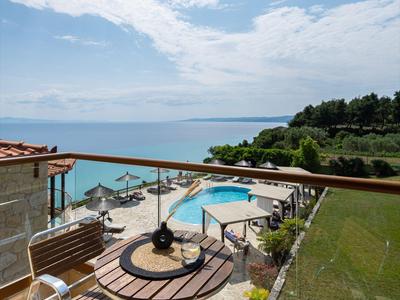 Balcon avec table et chaises donnant sur une piscine, la mer et un paysage boisé sous un ciel bleu.