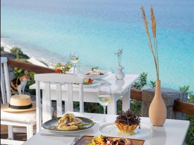 Stůl s jídlem na terase s výhledem na modré moře a pláž v pozadí.