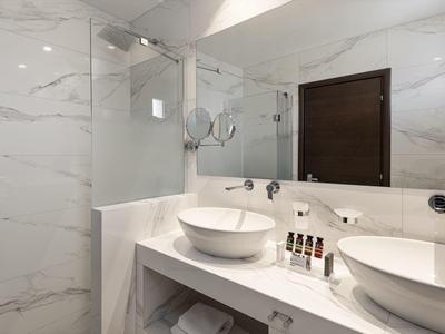 Moderní hotelová koupelna s bílými mramorovými stěnami, dvojitými umyvadly a toaletními potřebami.