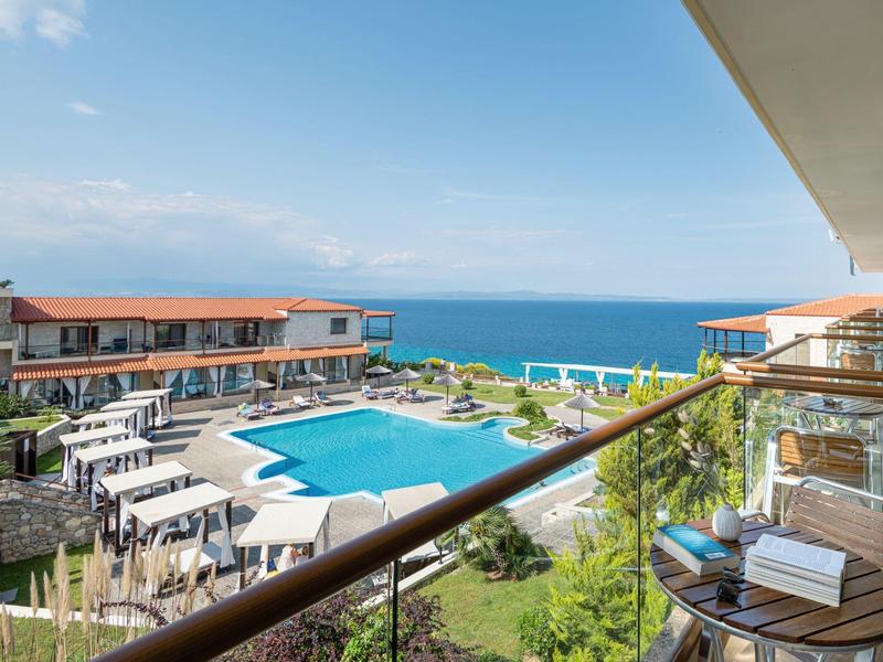 Balkon s stolem a židlemi s výhledem na bazén, hotelové budovy a moře v pozadí.