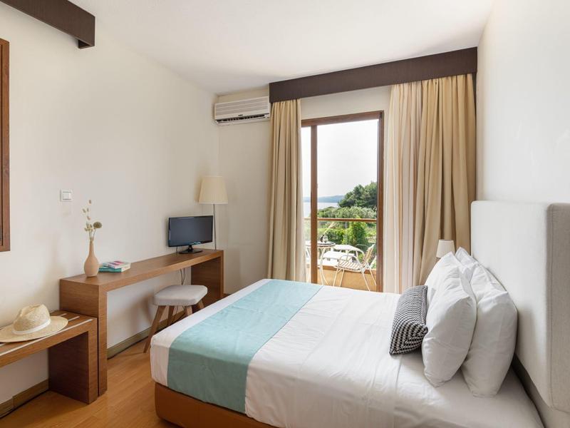 Jasný hotelový pokoj s manželskou postelí, TV, dřevěným nábytkem a výhledem na terasu.