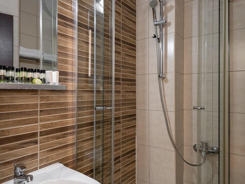 Malý sprchový kout se skleněnými dveřmi, bílým umyvadlem a hnědými obklady v hotelové koupelně.