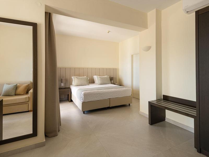 Světý hotelový pokoj s manželskou postelí, velkým zrcadlem a moderní skříní v neutrálních tónech.