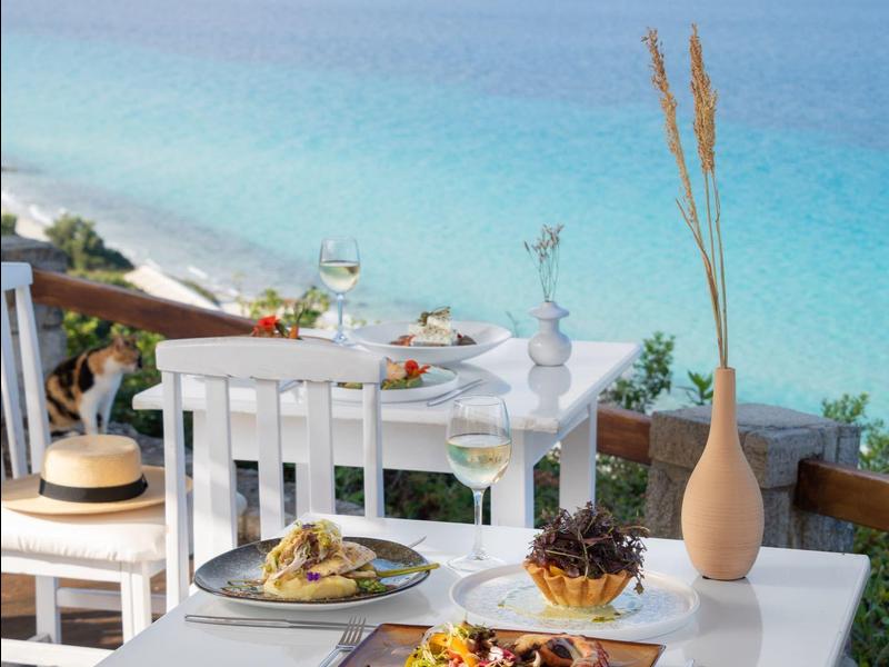 Stůl s jídlem na terase s výhledem na modré moře a pláž v pozadí.