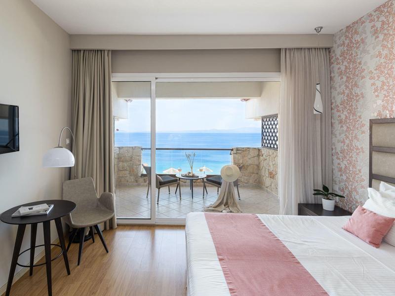 Světly hotelový pokoj s výhledem na moře, manželskou postelí a posezením na balkóně.
