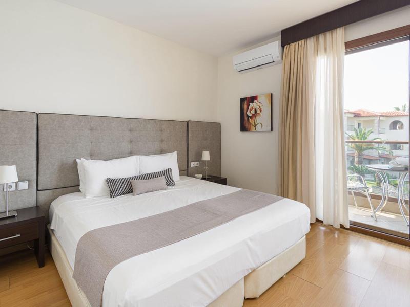 Světý hotelový pokoj s velkou postelí, nočními stolky a přístupem na balkon přes posuvné skleněné dveře.