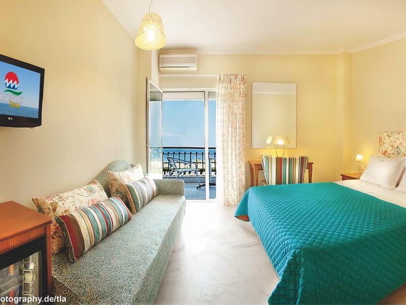 Camera d'albergo luminosa con balcone, divano, letto matrimoniale e TV a muro.