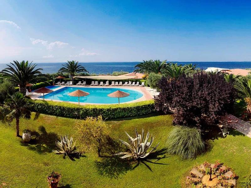 Giardino dell'hotel con piscina, lettini, palme e vista mare con cielo sereno.