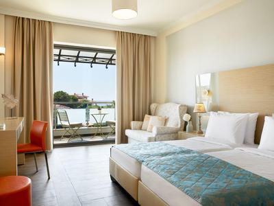Helles Hotelzimmer mit Doppelbett, Stuhl, Teppich, Balkon und See im Hintergrund.
