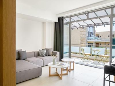 Helles Wohnzimmer mit grauem Ecksofa, zwei Holztischen und großem Fenster zum Balkon mit Stühlen.