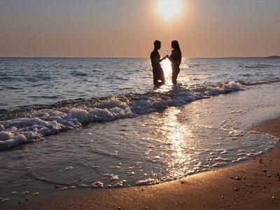 Ein Paar steht im Wasser am Strand, Sonnenuntergang im Hintergrund, warme Farben, sanfte Wellen.