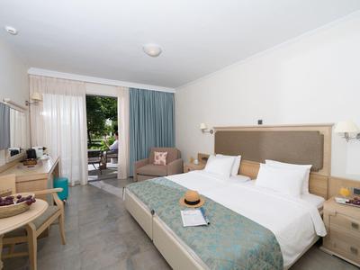 Moderní hotelový pokoj s manželskou postelí, pracovním stolem, křeslem a balkonem s výhledem do zeleně.