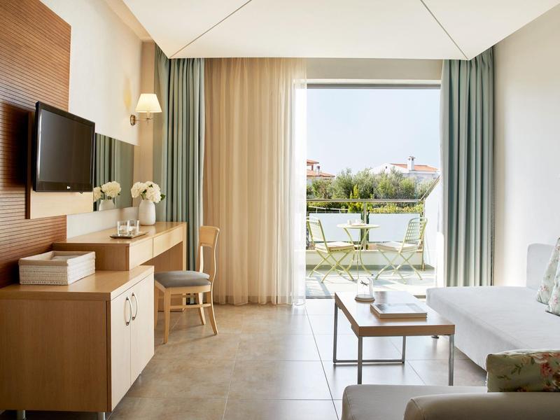 Světlo hotelový pokoj s moderním dřevěným nábytkem, pohovkou, televizí a výhledem na balkon.