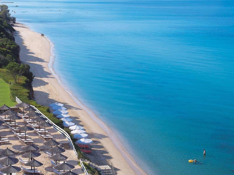 Dlouhá písčitá pláž s deštníky a čistým modrým mořem za slunečného počasí.