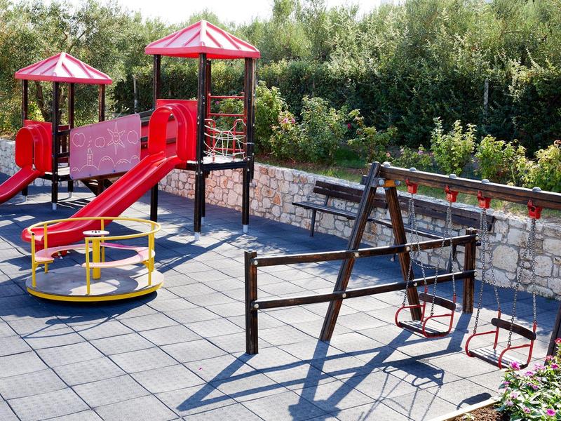 Dětské hřiště s houpačkami, skluzavkou a kolotočem vedle kamenné zdi.
