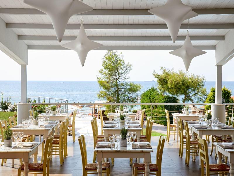 Otevřená restaurace s výhledem na moře, bílým nábytkem a zelenou zahradní výzdobou.