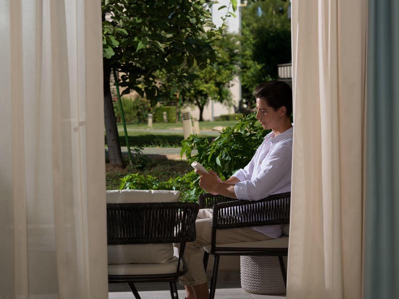 Osoba sedí uvolněně na židli na balkóně s výhledem na zelenou zahradu