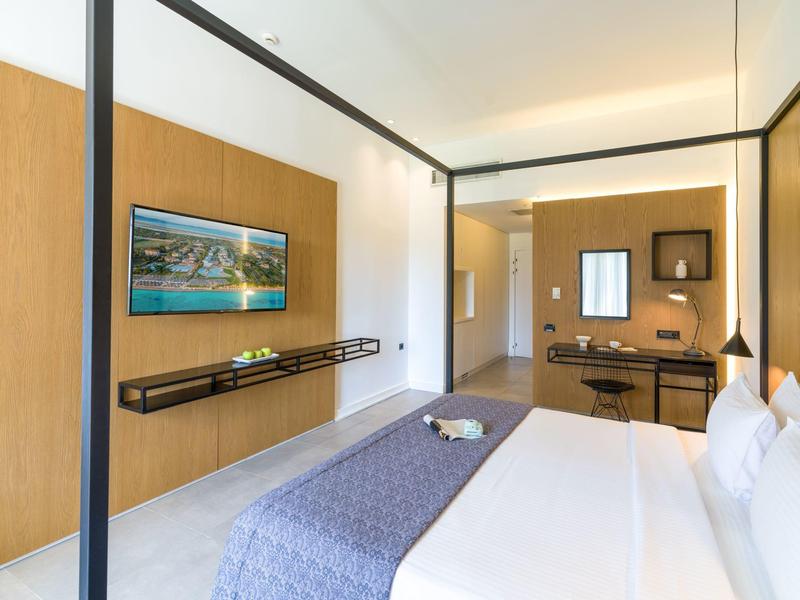 Moderně zařízený hotelový pokoj s manželskou postelí, nástěnnou TV a psacím stolem v světlém prostředí.