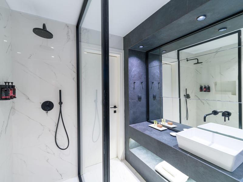 Moderní hotelová koupelna s velkým zrcadlem, bílým umyvadlem a dešťovou sprchou s mramorovým vzhledem.