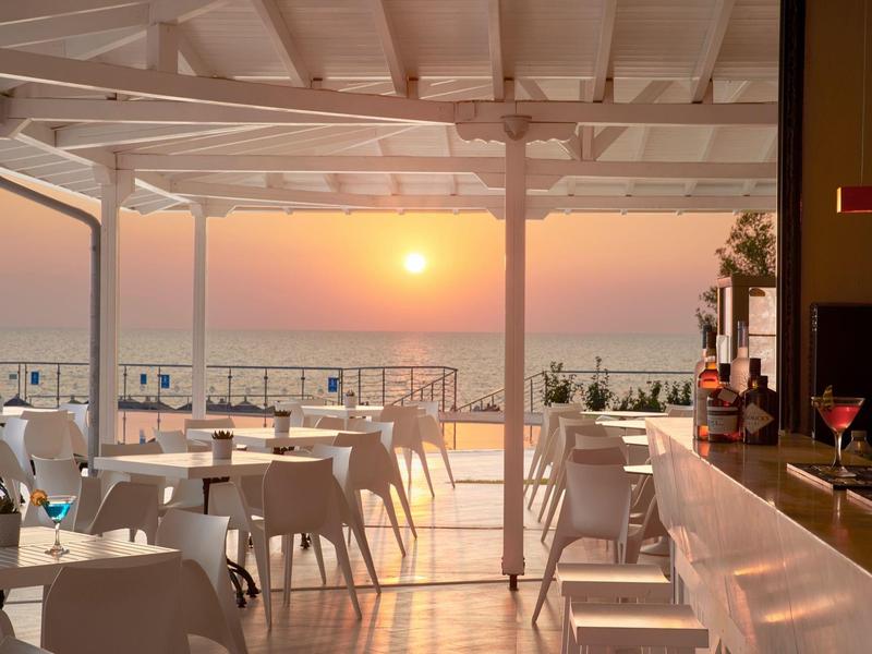 Otevřená terasa s bílými stoly a židlemi při západu slunce u moře.