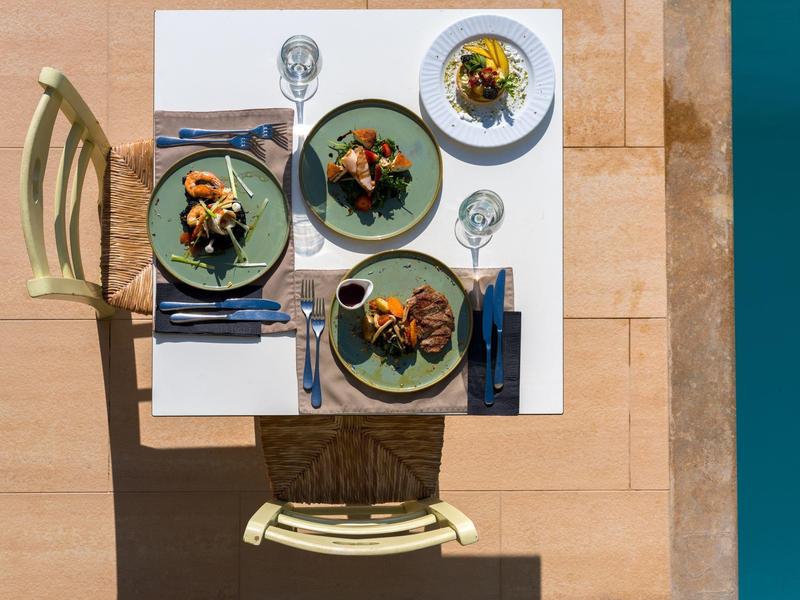 Pohled shora na prostřený stůl se třemi talíři a příbory na terase u vody.