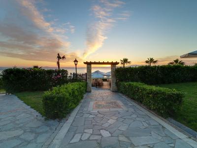 Ein gepflasterter Weg führt durch einen Garten zu einem Tor mit Blick auf das Meer bei Sonnenuntergang.