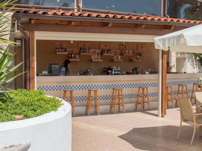 Offene Bar mit Holzstühlen neben einer Sonnenterrasse in einem Hotelgarten.