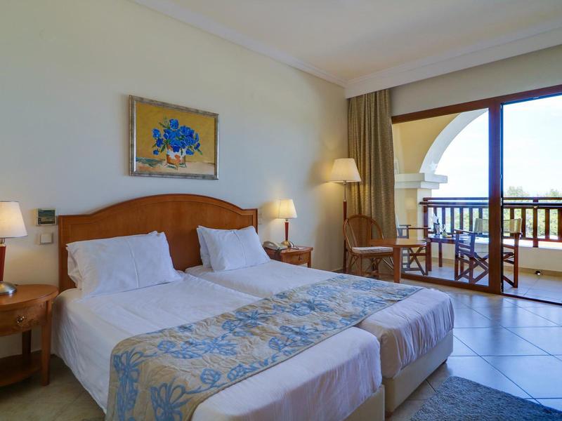 Hotelový pokoj se dvěma jednolůžky, nočními stolky a balkonem s výhledem ven.