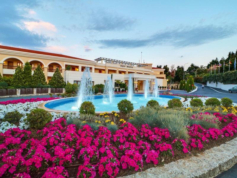 Hotelový komplex s fontánou a kvetoucími záhony při západu slunce.