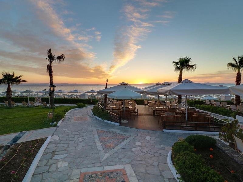 Západ slunce na terase s stoly, slunečníky a palmami u vody.