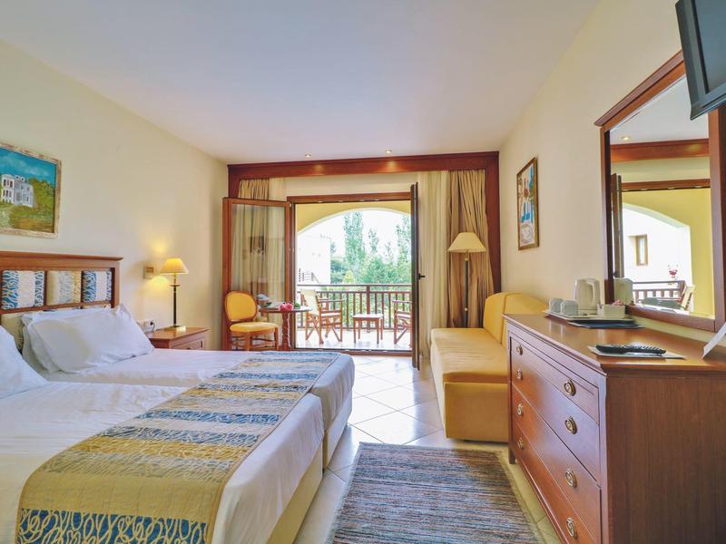 Světelný hotelový pokoj s manželskou postelí, balkonem a dřevěným nábytkem v teplých tónech.