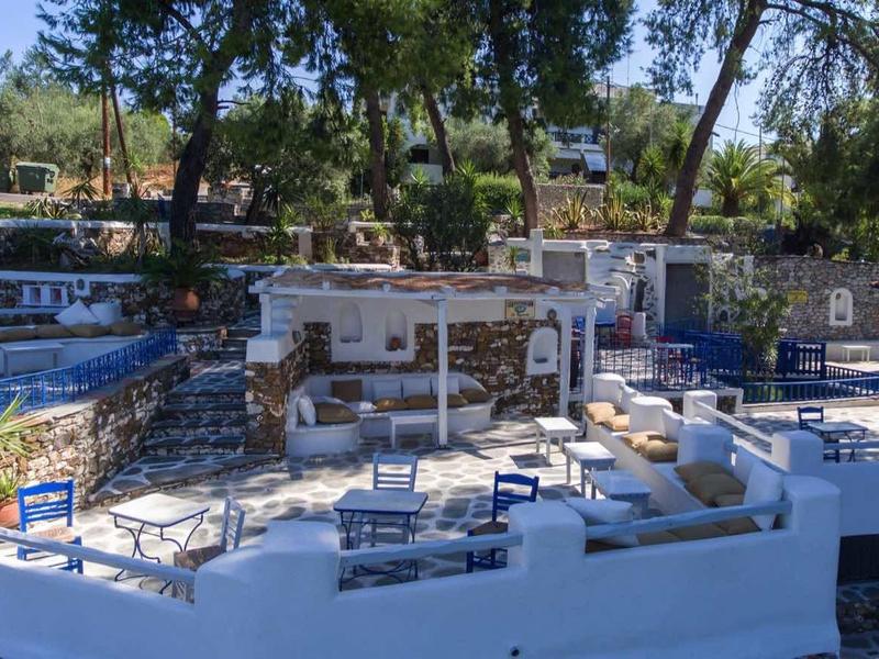 Taras na zewnątrz z białymi ścianami, stołami, ławkami i drzewami w słonecznym otoczeniu.