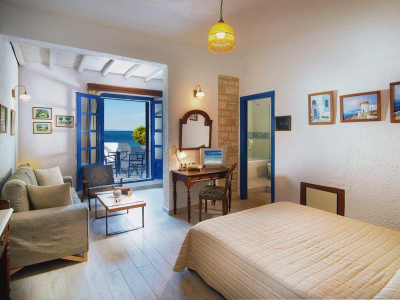 Przytulny pokój hotelowy z łóżkiem, sofą, biurkiem i otwartymi drzwiami balkonowymi z widokiem na morze.