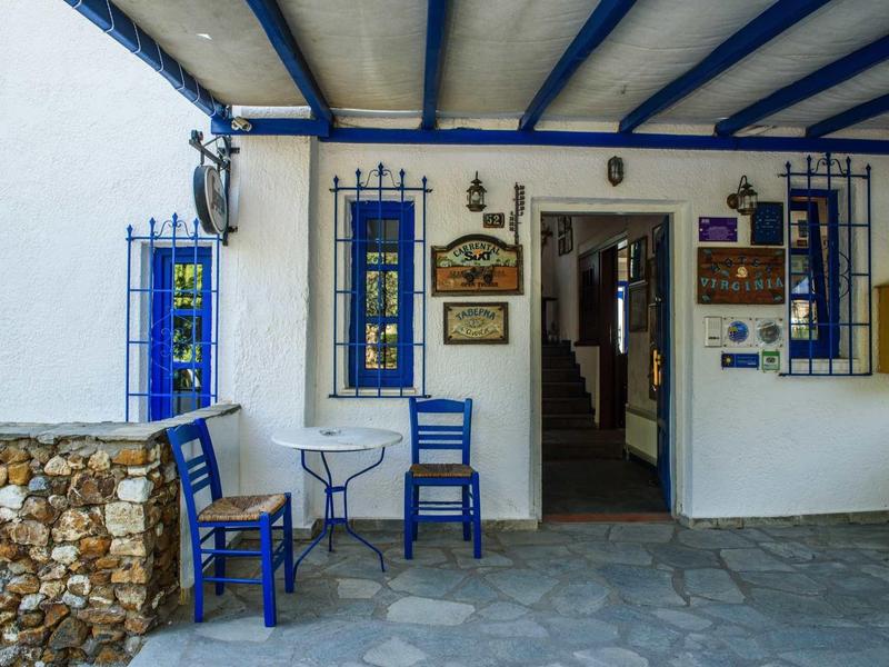 Przytulne miejsce na zewnątrz z niebieskimi krzesłami i stolikiem pod niebieskimi belkami przy białym wejściu.