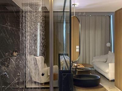 Moderní hotelový pokoj se skleněnou sprchou, pohodlným křeslem a bílou postelí.