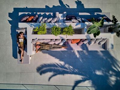 Vista aérea de una zona de piscina soleada con mesas largas y bancos en el jardín de un hotel.