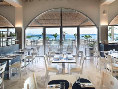 Restaurante luminoso con sillas y mesas blancas, gran ventana arqueada con vista al mar.