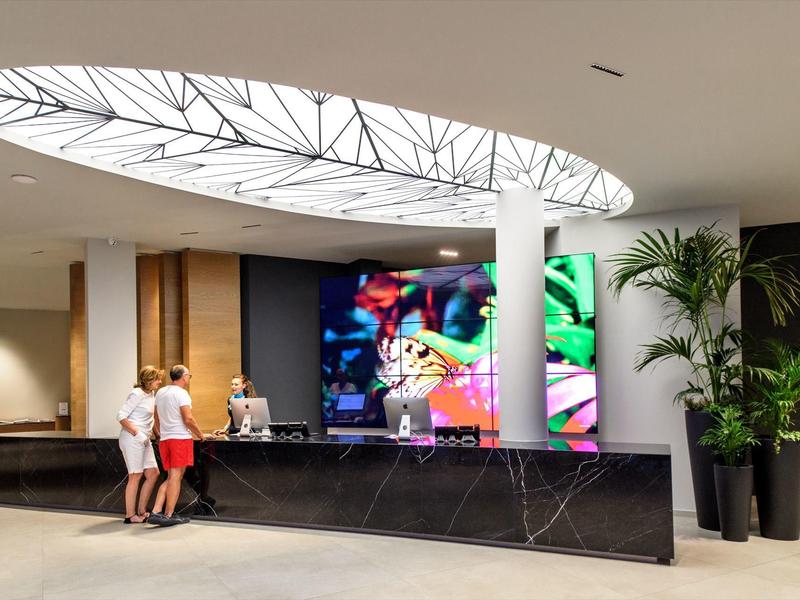 Moderní hotelová lobby s černou recepcí, velkými rostlinami a digitálním uměleckým dílem na zdi