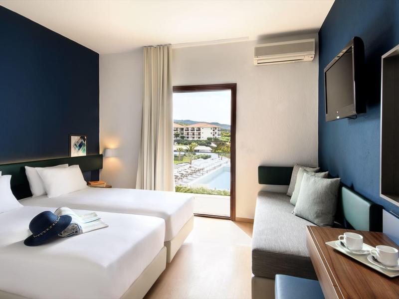 Moderní hotelový pokoj se dvěma postelemi, pohovkou, psacím stolem a výhledem na venkovní prostor.